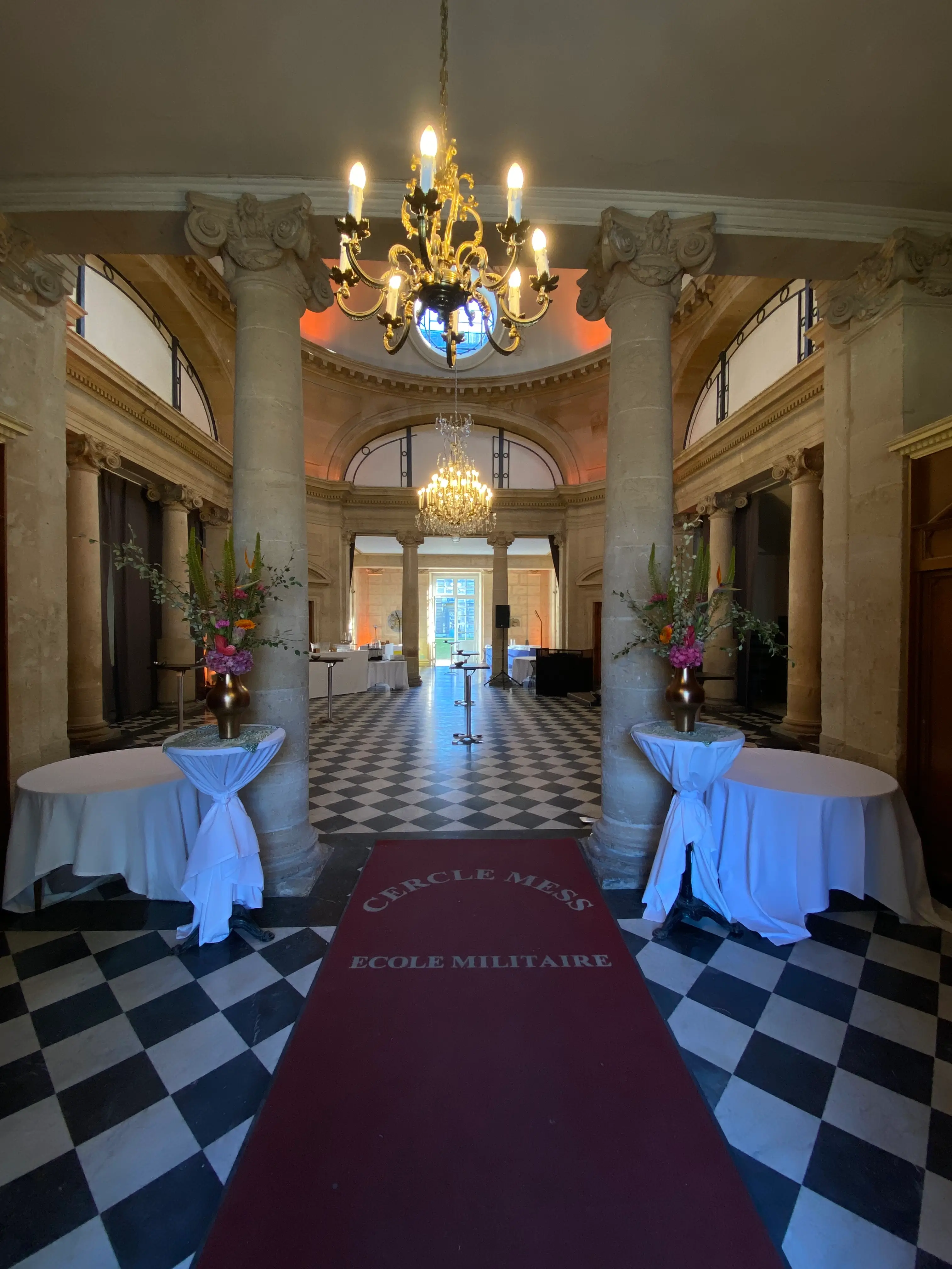 Hall à colonnes et lustre du Cercle-Mess de l'École militaire, tapis rouge et mange-debout dressés pour un bal d'officiers